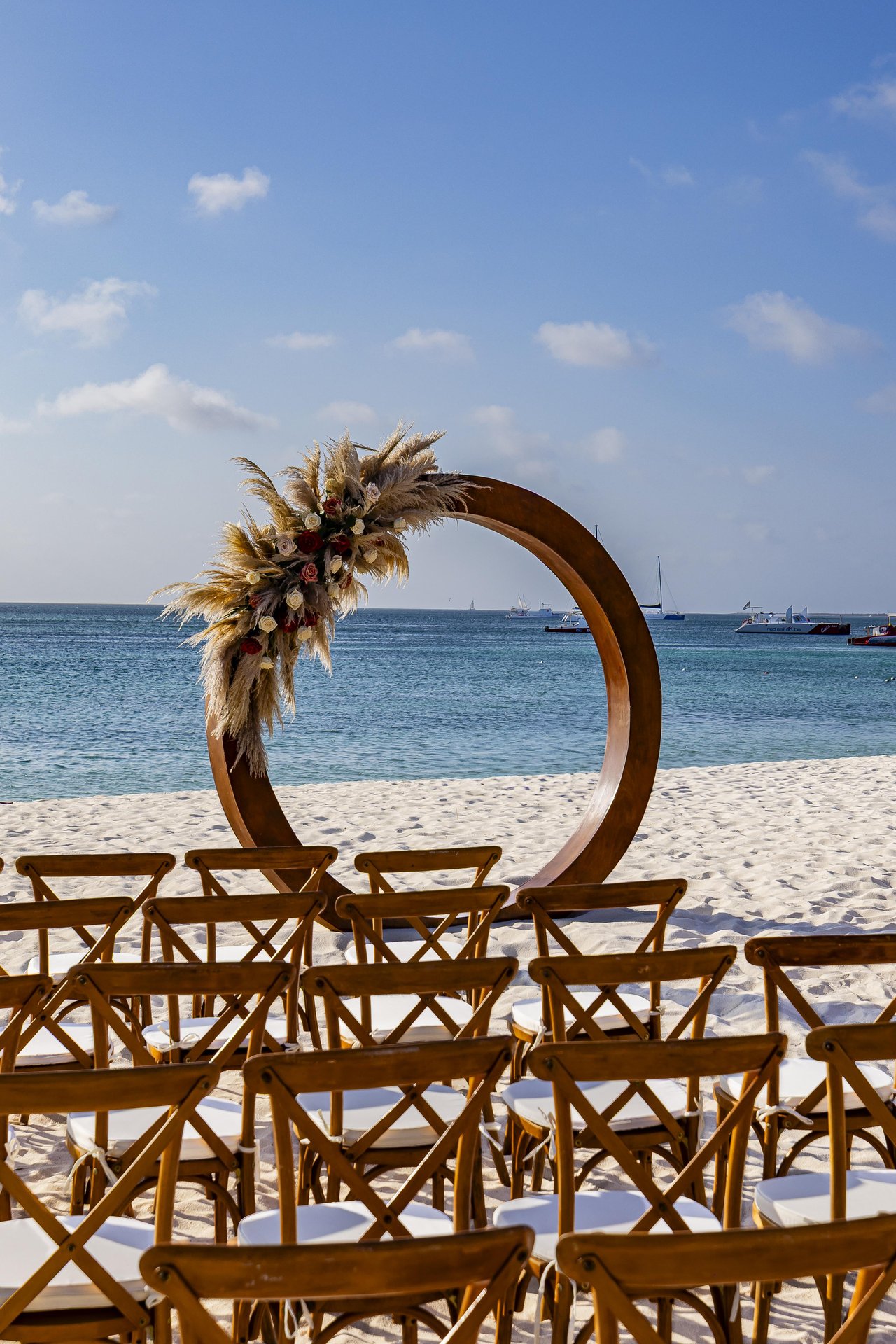 Wooden chairs are arranged on a sandy beach facing a large circular wooden arch decorated with pampas grass and flowers, with the ocean and boats in the background under a blue sky.