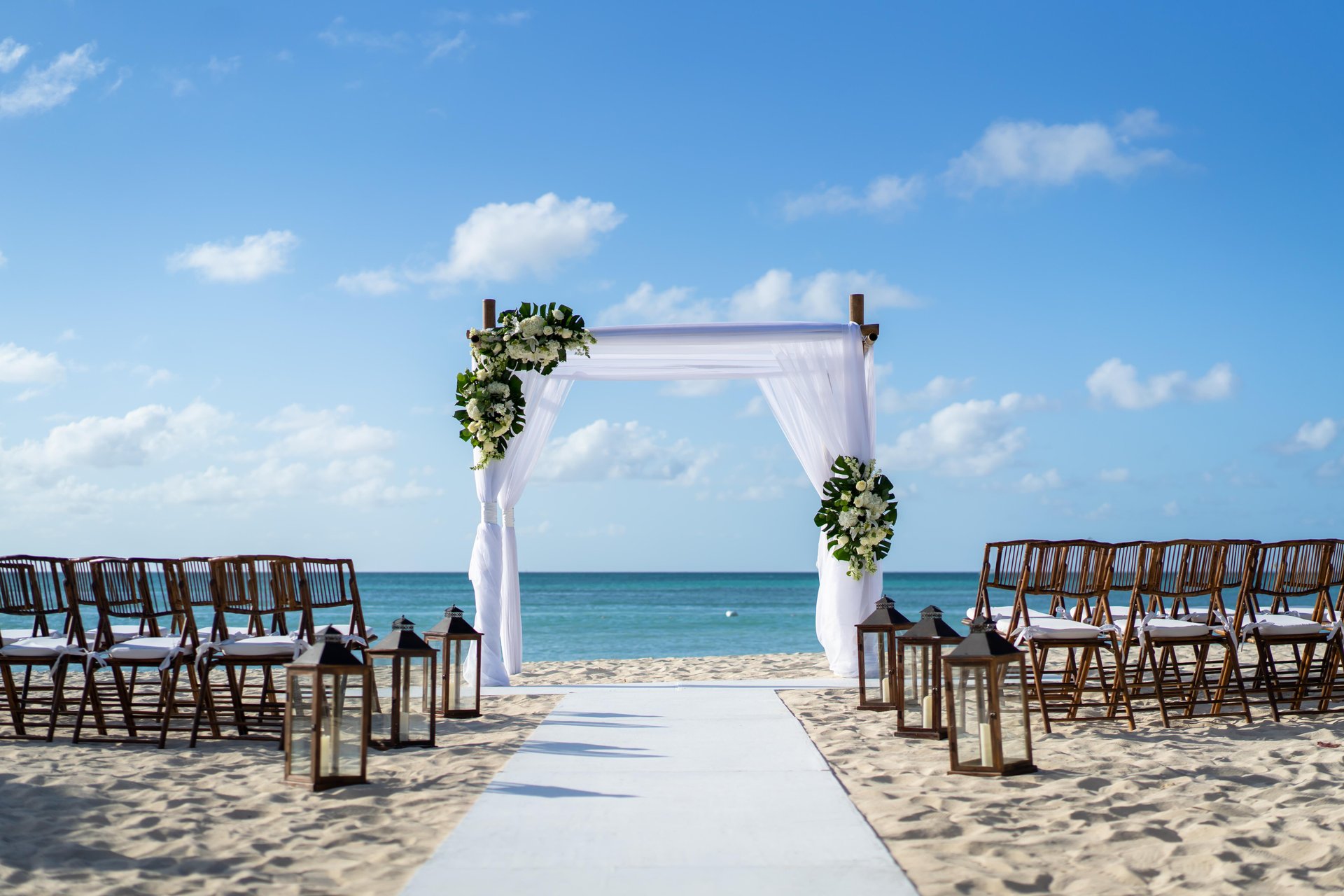 A beach wedding setup with an aisle leading to a white arch decorated with greenery and flowers, surrounded by wooden chairs and lanterns, overlooking the ocean under a blue sky with scattered clouds.