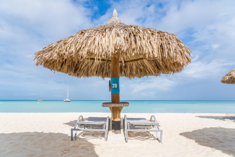 A thatched beach umbrella with two lounge chairs underneath sits on white sand facing turquoise ocean water, with a sailboat in the distance under a partly cloudy sky.