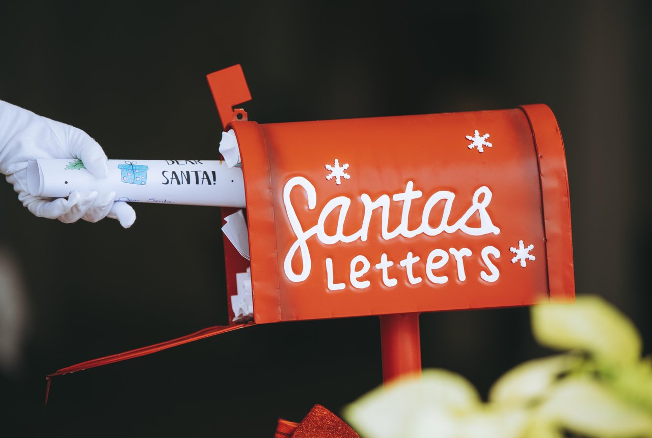 A hand puts a letter into a red mailbox labeled "Santa's Letters," decorated with white snowflakes, against a dark background.