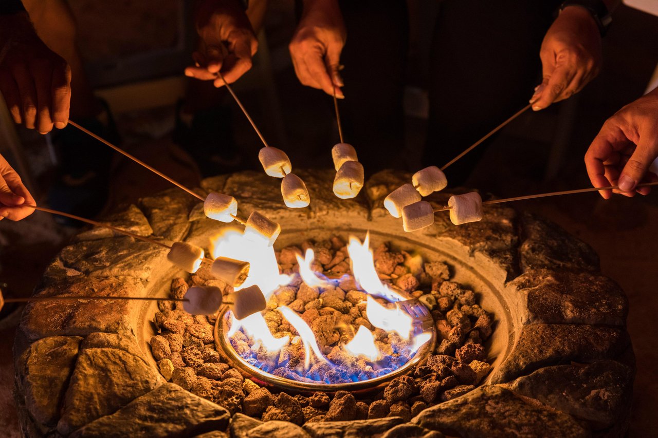 Several people toast marshmallows on skewers over an outdoor stone fire pit with bright flames, suggesting a social gathering or campfire activity at night.