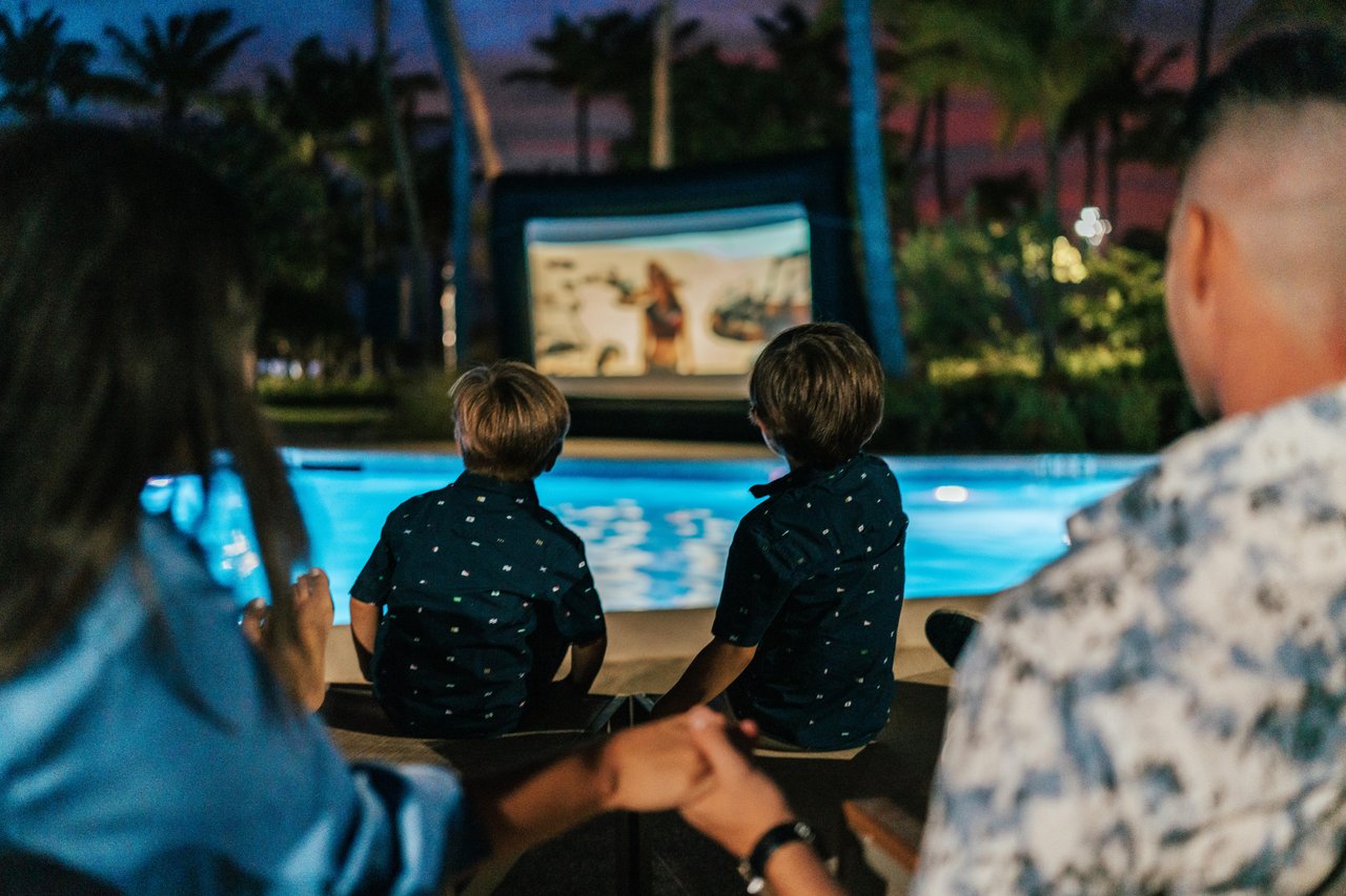 A family sits together beside a pool at night, watching a movie projected on an outdoor screen surrounded by palm trees. Two children, seen from behind, are focused on the movie as adults sit nearby.