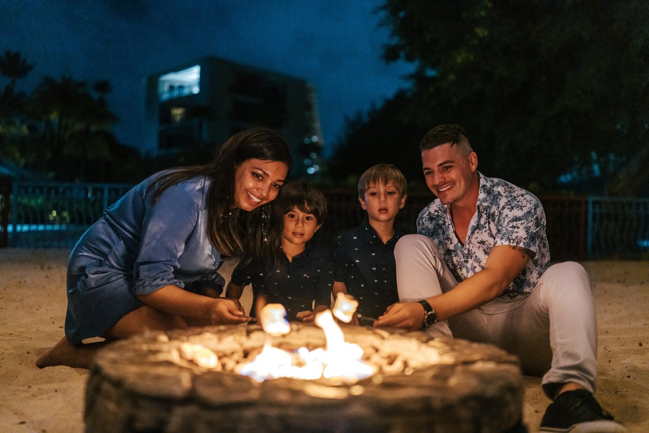 A smiling family of four sits on sand around a campfire at night, roasting marshmallows. There are trees and a building in the background, and everyone appears happy and relaxed.