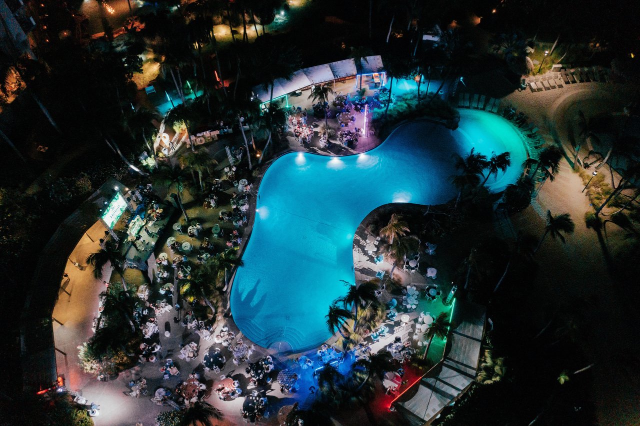 Aerial view of a large, curvy swimming pool at night, surrounded by palm trees and outdoor dining tables, all lit with colorful lights in blue, green, and purple hues.