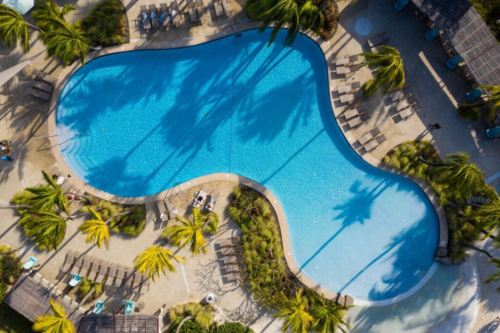 Aerial view of a large, curvy swimming pool surrounded by palm trees, lounge chairs, and people relaxing in the sun at a resort. The bright blue water contrasts with the tan pool deck.