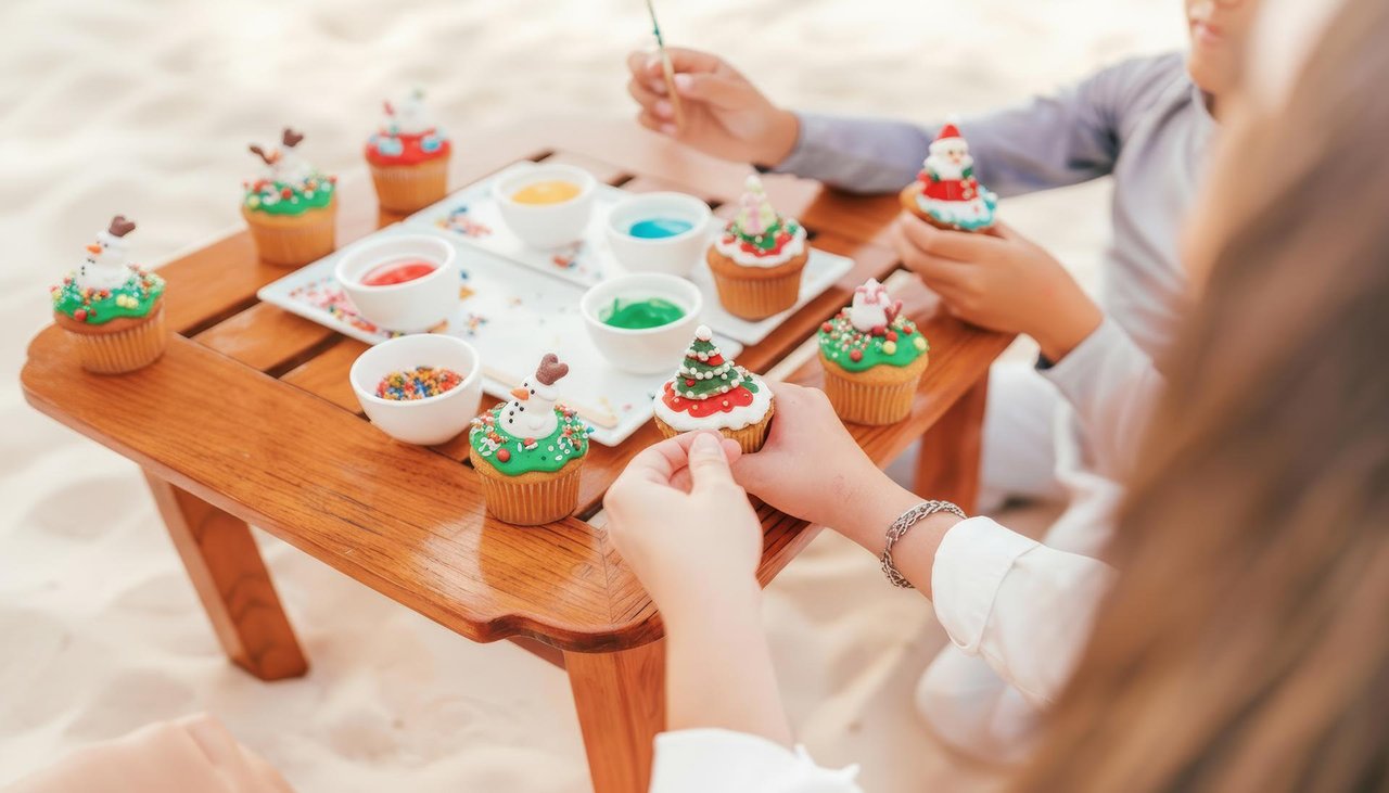 Children decorate cupcakes with colorful icing and toppings on a small wooden table. The cupcakes have festive designs, and bowls of sprinkles and icing are arranged in the center. The setting appears to be outdoors.