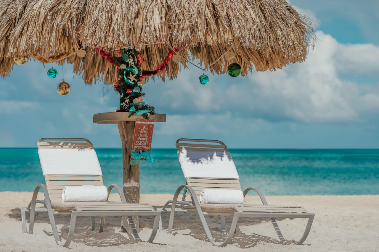 Two empty lounge chairs with white towels sit on sandy beach under a thatched umbrella decorated with Christmas ornaments, facing clear turquoise ocean and a blue sky with fluffy clouds.
