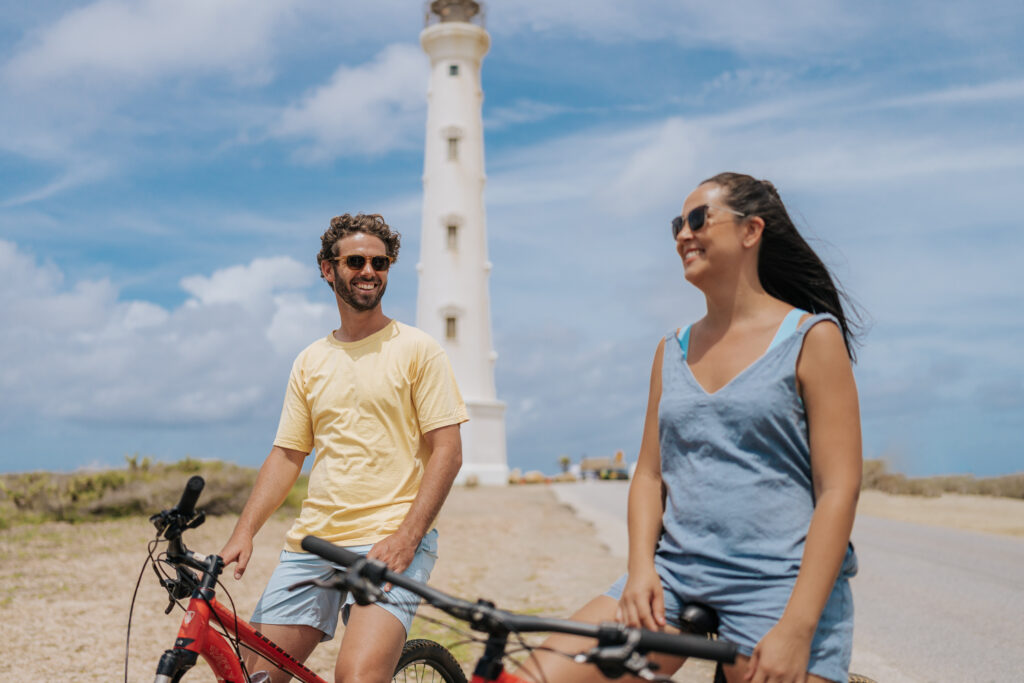 Two people wearing sunglasses ride bikes and smile near a tall white lighthouse on a sunny day, with blue sky and some clouds in the background.