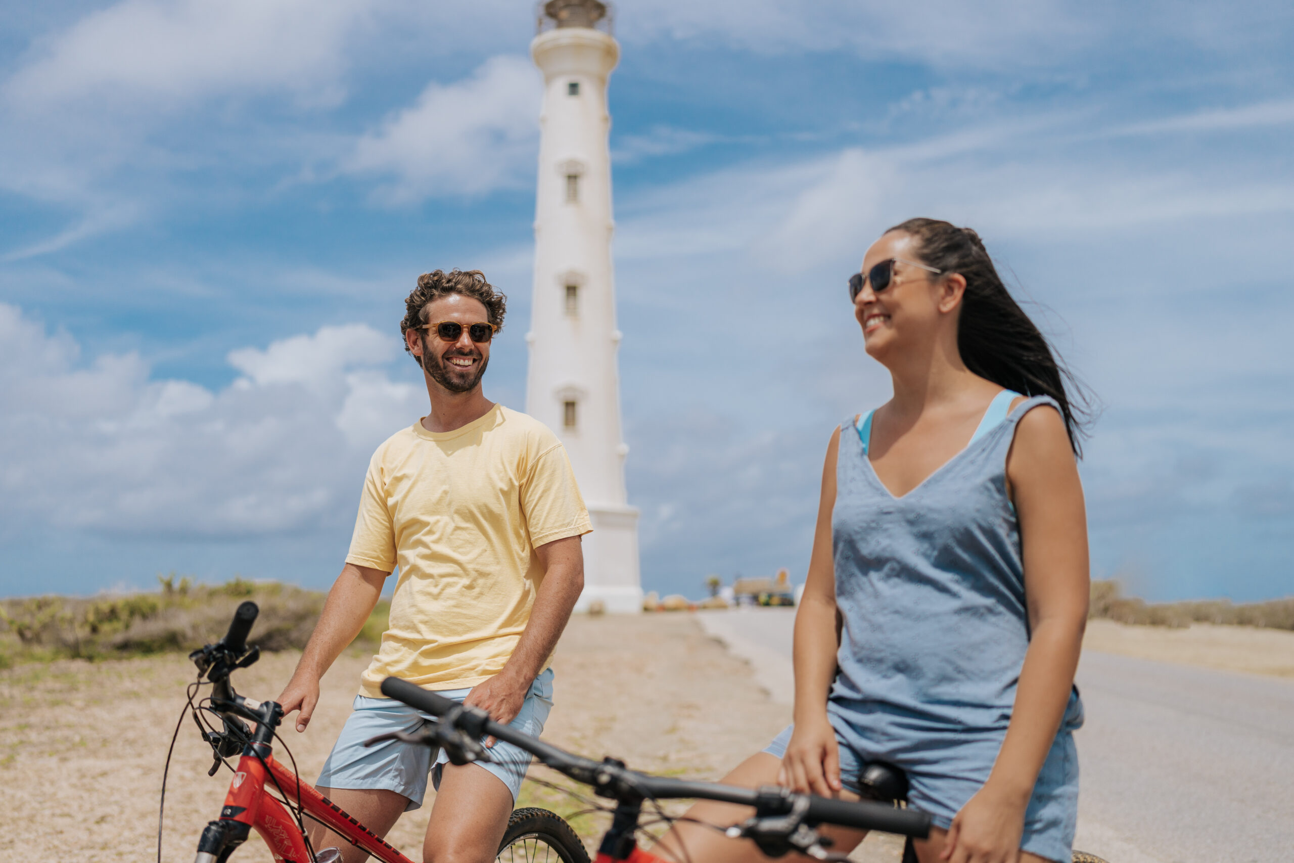 Two people wearing sunglasses ride bikes and smile near a tall white lighthouse on a sunny day, with blue sky and some clouds in the background.