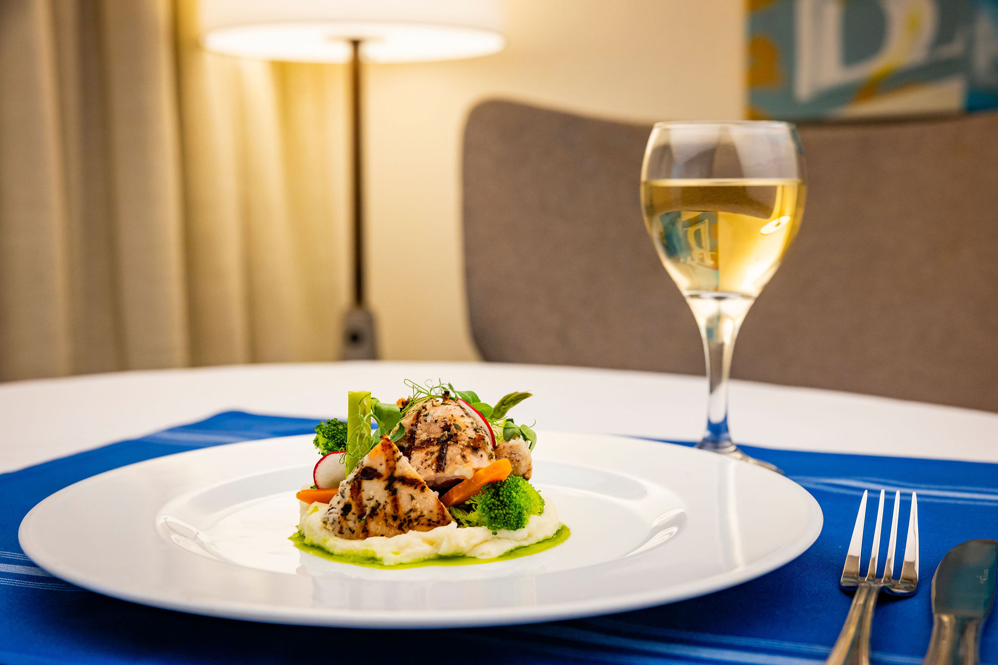 A plated gourmet meal featuring grilled chicken on a bed of mashed potatoes with assorted vegetables, set on a blue placemat next to a glass of white wine, with a modern lamp and chair in the background.