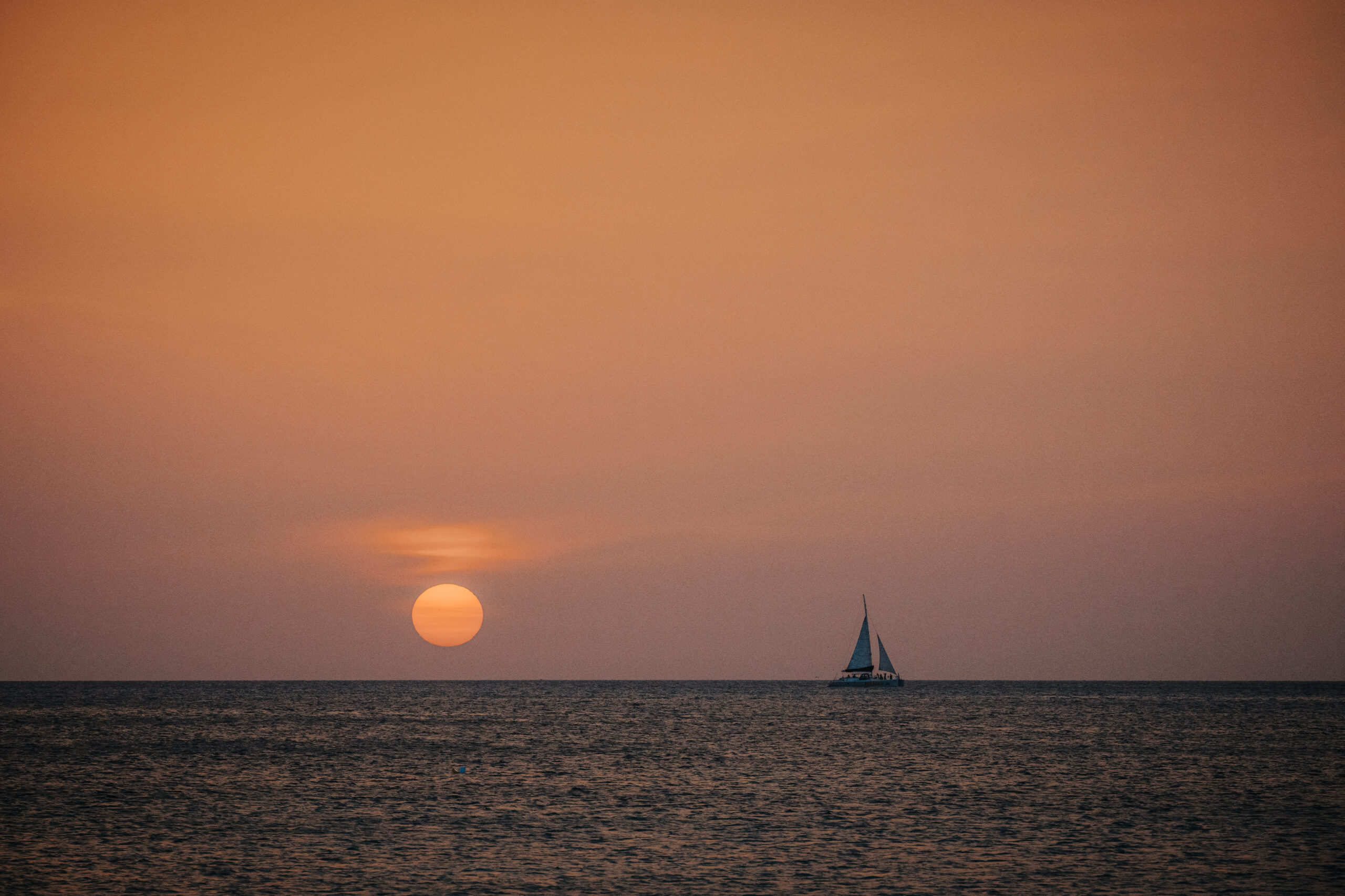 A sailboat floats on calm ocean waters during sunset, with the sun low on the horizon and the sky glowing in warm orange tones.