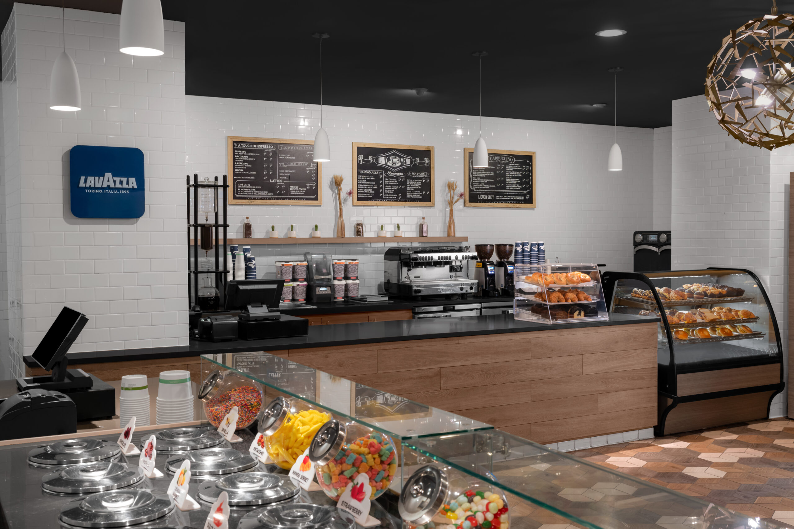 A modern café interior with a Lavazza sign, coffee machines, menu boards on the wall, display cases filled with pastries, and jars of colorful candies on the counter. The space has pendant lights and a clean, inviting look.