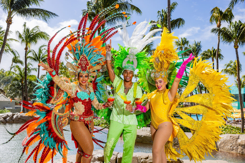 Three people in vibrant carnival costumes—red, green, and yellow—stand outdoors near palm trees and water, smiling and holding drinks, celebrating in a festive, tropical setting.
