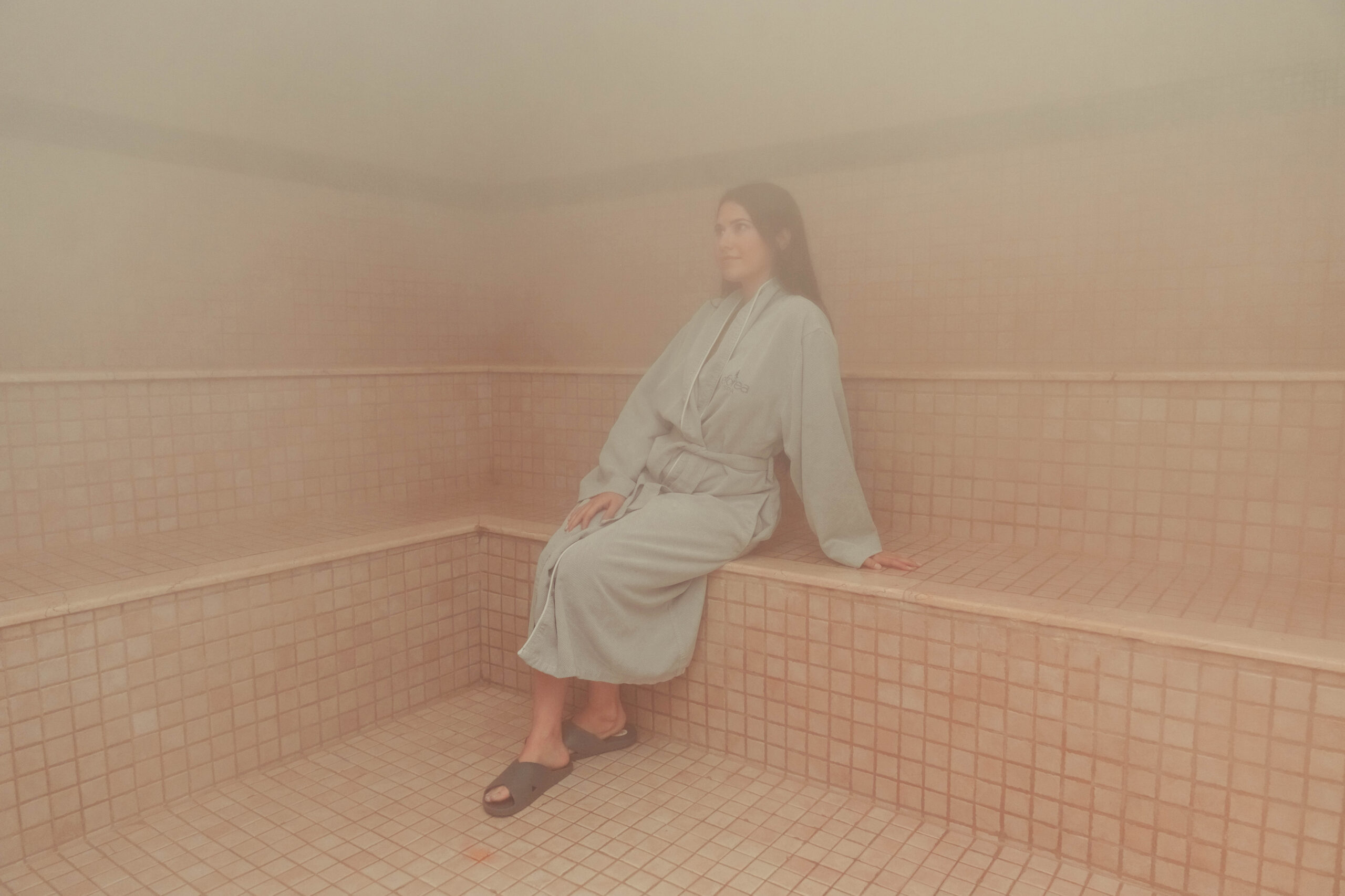A woman in a light gray robe sits on tiled benches inside a steam room, surrounded by thick mist. She rests one hand on the bench and looks ahead, appearing relaxed.
