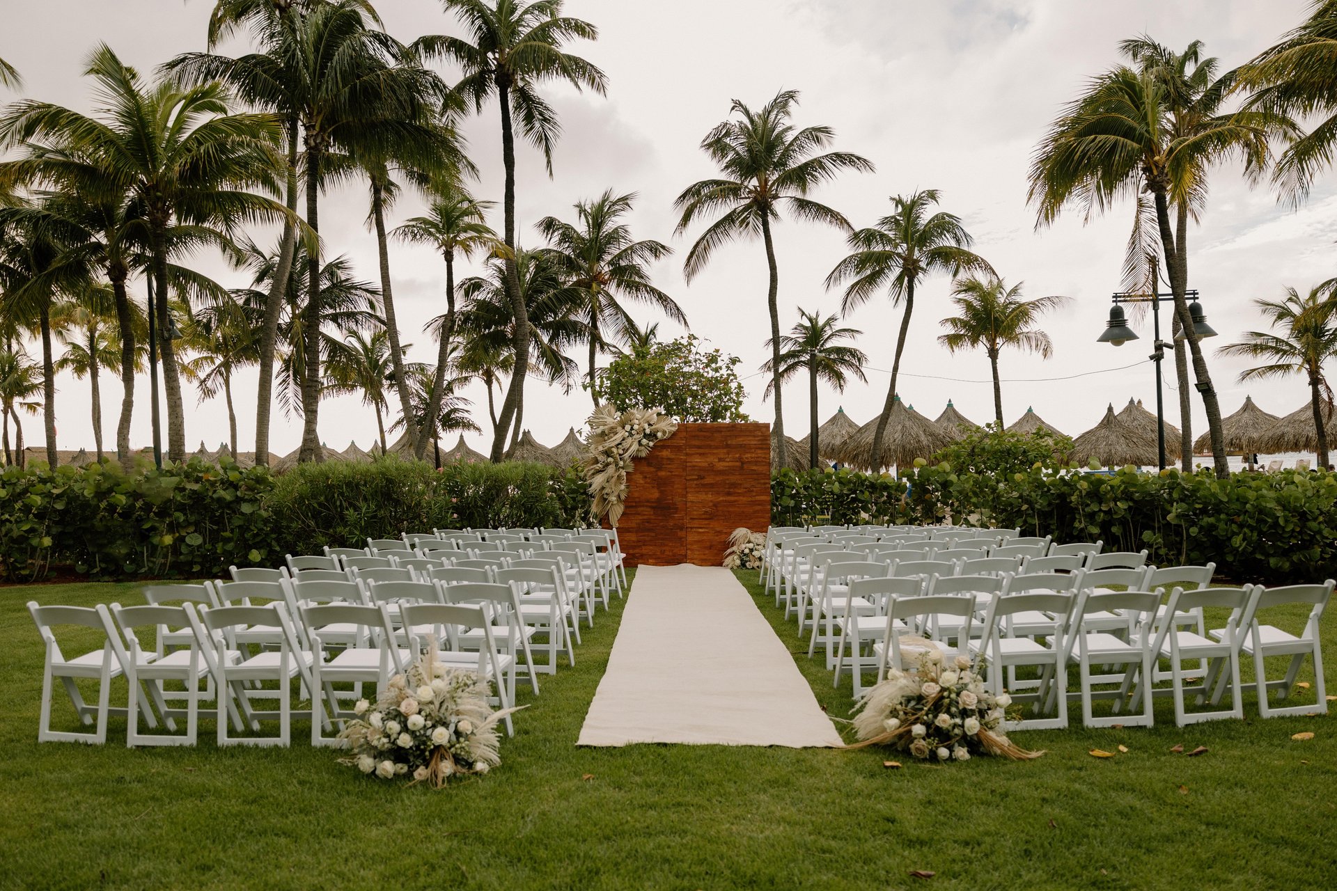 An outdoor wedding setup with rows of white chairs on green grass, a white aisle runner, floral arrangements, palm trees in the background, and a wooden backdrop at the ceremony site.