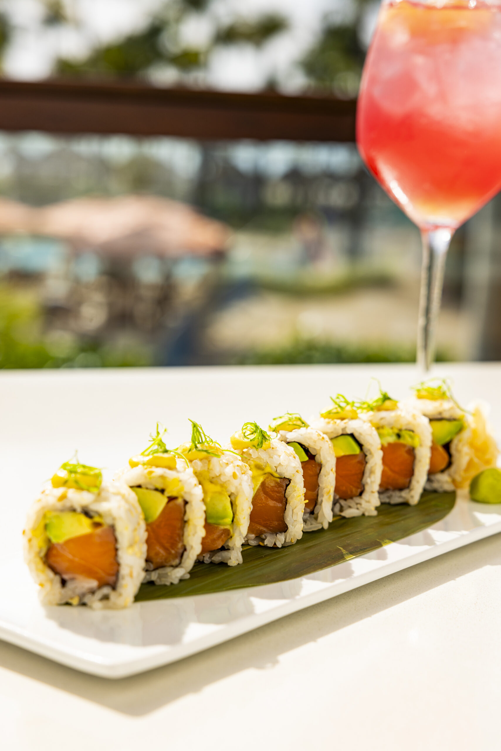 A plate of sushi rolls filled with salmon and avocado, garnished with herbs, sits on a white table next to a glass of pink drink with ice; an outdoor setting is blurred in the background.