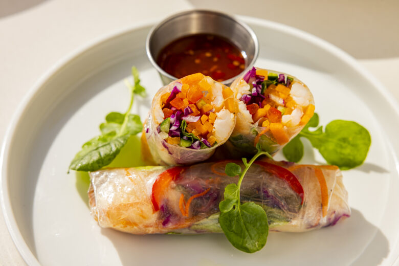 Three fresh vegetable spring rolls, filled with colorful vegetables and shrimp, are served on a white plate with leafy greens and a small metal cup of dipping sauce.
