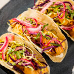Three tacos filled with crispy fried fish, shredded cabbage, sliced red onion, cilantro, and creamy orange sauce are arranged on a dark plate with lime wedges on the side.