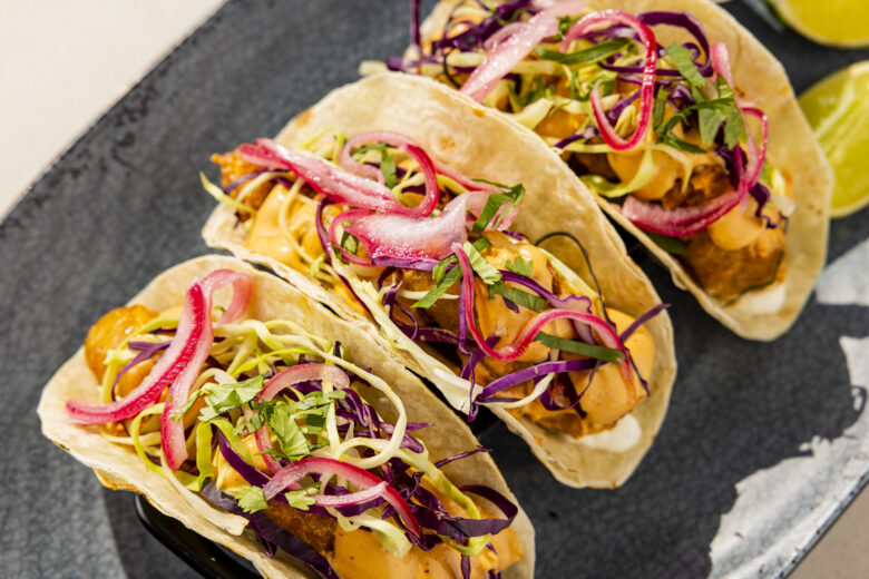 Three tacos filled with crispy fried fish, shredded cabbage, sliced red onion, cilantro, and creamy orange sauce are arranged on a dark plate with lime wedges on the side.