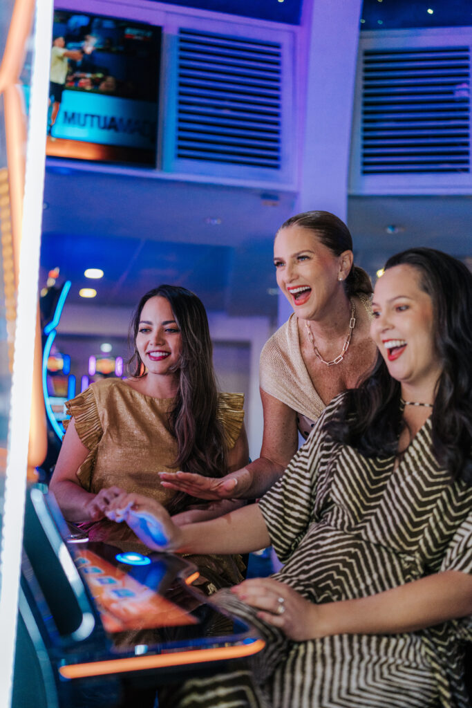 Three women smiling and playing a slot machine in a brightly lit casino. Two are standing while one is seated at the machine, all appearing to enjoy the game together.