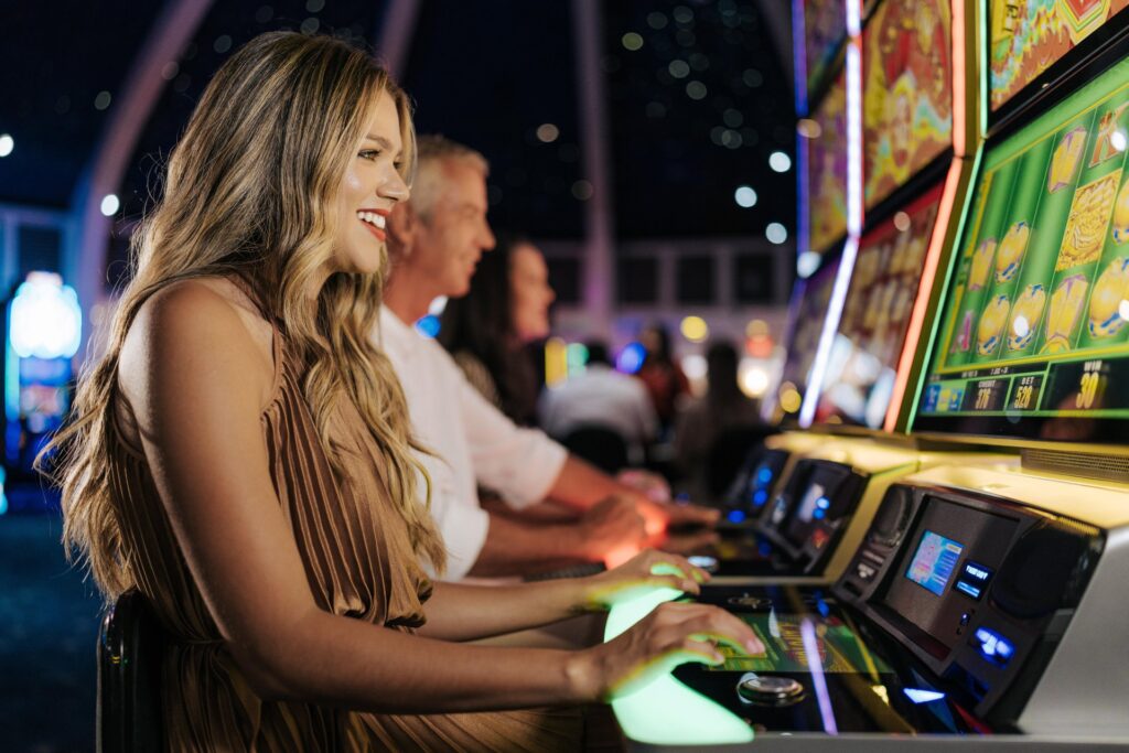 A young woman smiles while playing a brightly lit slot machine in a casino, with two other people sitting beside her and enjoying the games.