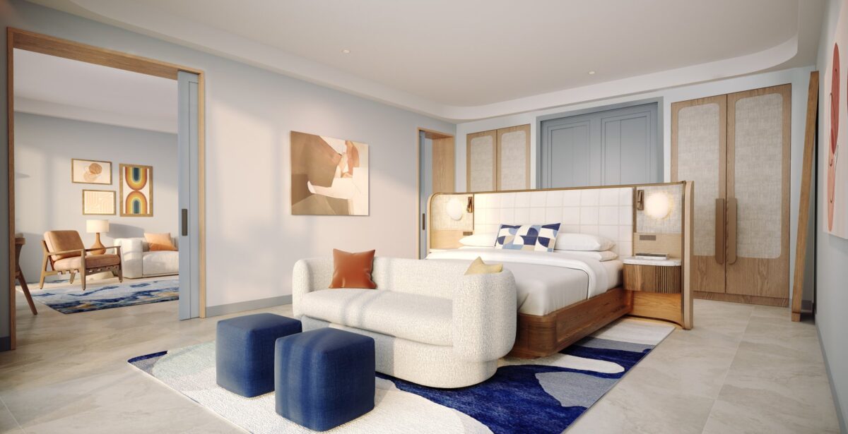 A modern hotel room features a king bed with a padded headboard, a white sofa, two blue ottomans, abstract art, wood accents, and an adjacent sitting area with a rug and armchair visible through sliding doors.