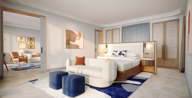 A modern hotel room features a king bed with a padded headboard, a white sofa, two blue ottomans, abstract art, wood accents, and an adjacent sitting area with a rug and armchair visible through sliding doors.