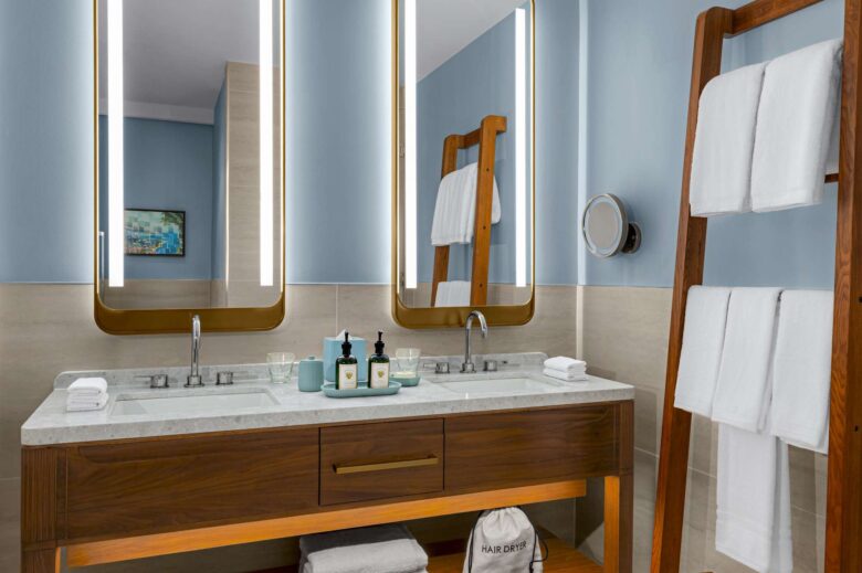 Modern bathroom with double sink vanity, two large mirrors with lights, a wooden towel ladder holding white towels, toiletries on the counter, and a hair dryer bag below. Walls are light blue with beige tile accents.