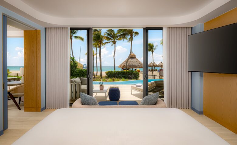 A hotel room with a bed facing sliding glass doors that open to a patio with lounge chairs, a pool, palm trees, and a view of the ocean under a sunny sky.