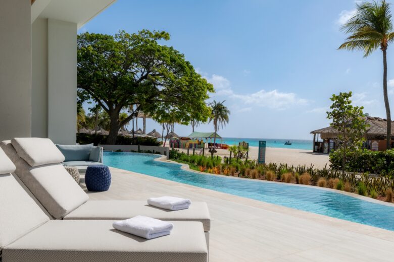 Two lounge chairs with towels sit beside a pool overlooking a sandy beach, palm trees, and the ocean under a clear blue sky.
