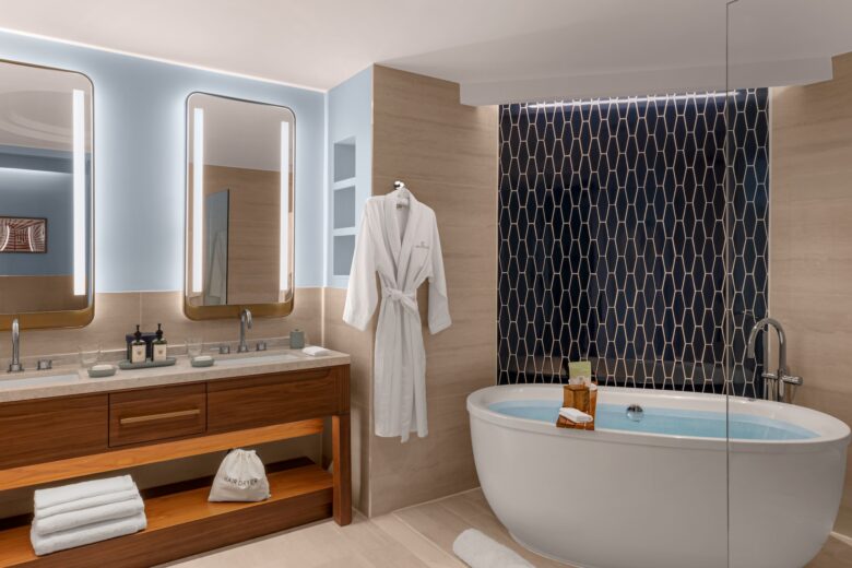Modern bathroom with a double sink vanity, two mirrors, a hanging white bathrobe, towels, and a freestanding bathtub filled with water, set against a dark geometric tile wall.