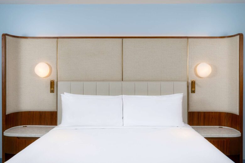 A neatly made bed with white linens, a padded headboard, and two round wall lamps on either side. The headboard and surrounding panels are beige with wood accents.