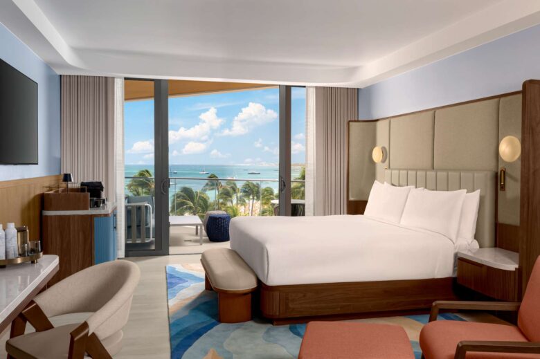 Modern hotel room with a large bed, seating area, TV, and floor-to-ceiling windows opening to a balcony with ocean and palm tree views under a blue sky with clouds.