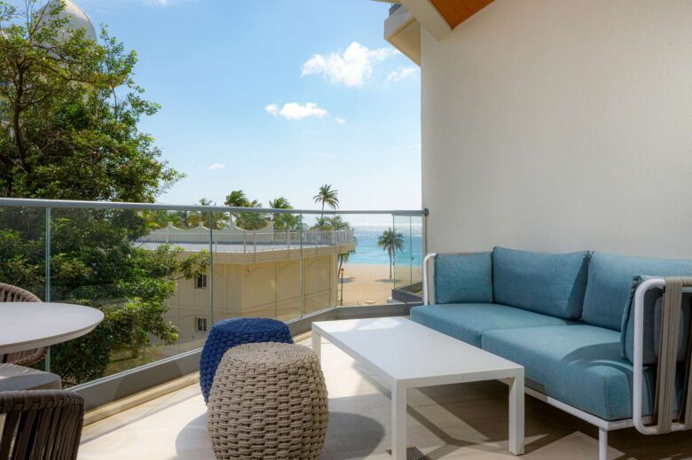 Modern balcony with blue sofa, round woven stools, and glass railing overlooking a sandy beach, palm trees, and the ocean under a bright, sunny sky.