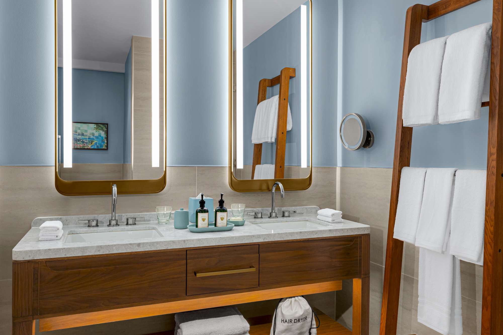 Modern hotel bathroom with a double sink vanity, lighted mirrors, wooden towel rack holding white towels, and toiletries neatly arranged on the counter. A labeled hair dryer bag sits on a shelf below the sink.