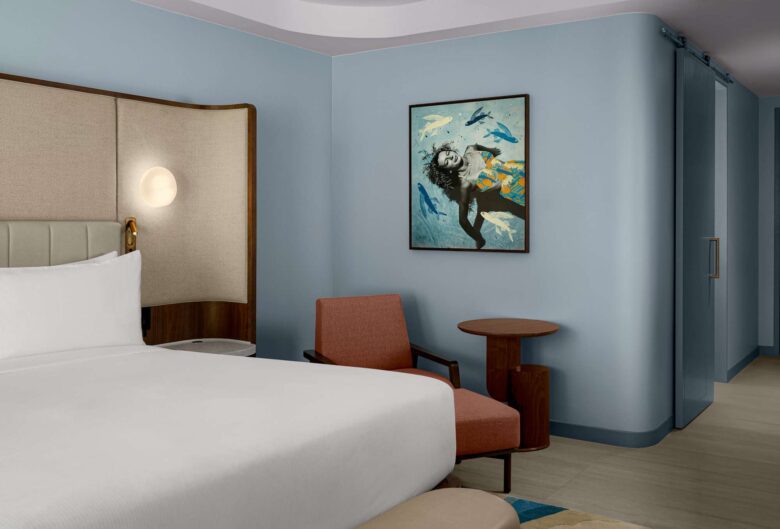 Modern hotel room with a neatly made bed, padded headboard, red armchair, round wooden tables, and a wall art featuring a person surrounded by birds. Walls are light blue, and there is a glass door in the background.