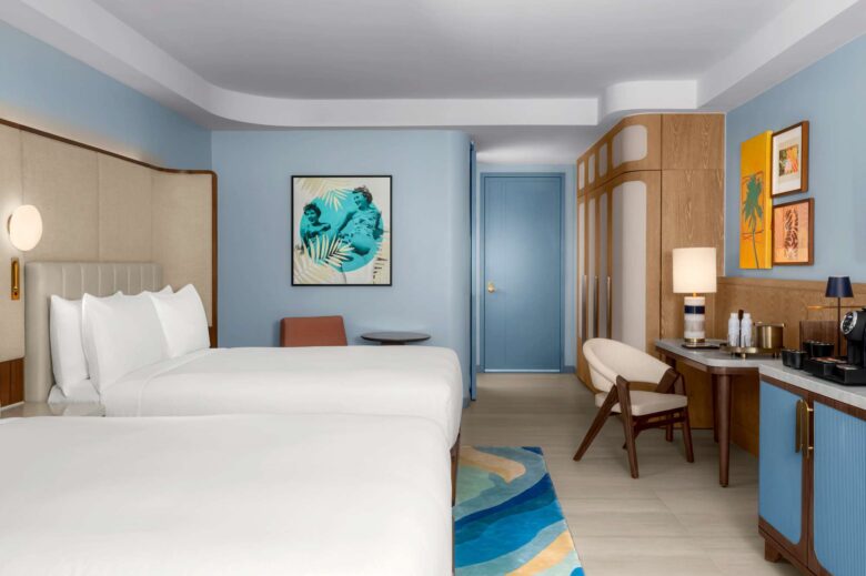 A modern hotel room with two double beds, blue accents, a desk, chair, wardrobe, minibar, wall art, and a lamp. The decor features soft lighting and a blue, beige, and wood color scheme.
