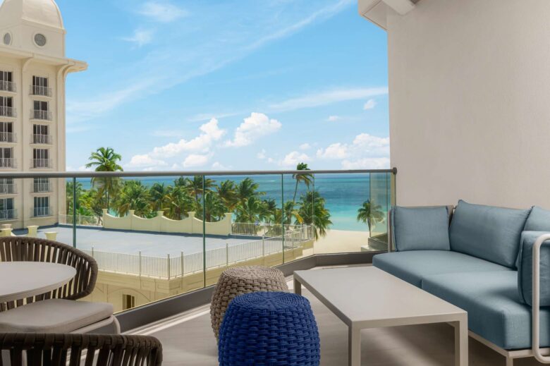 Balcony with modern outdoor furniture overlooking palm trees, a white building, and the ocean under a bright blue sky with scattered clouds.