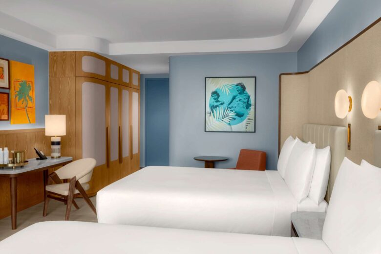 Modern hotel room with two white beds, light wood details, a desk and chair, an orange lounge chair, and colorful artwork on pale blue walls. Soft lighting creates a calm, inviting atmosphere.