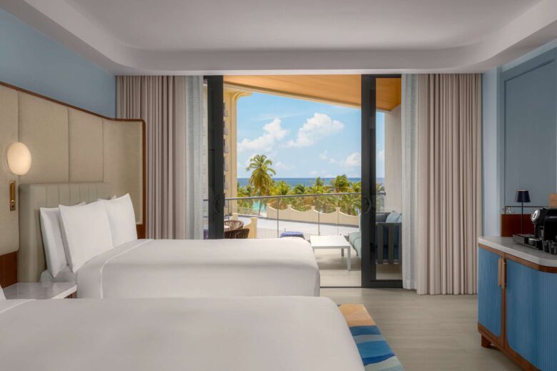 Hotel room with two beds, a coffee station, and large windows opening to a balcony with outdoor seating, palm trees, and an ocean view under a blue sky with scattered clouds.