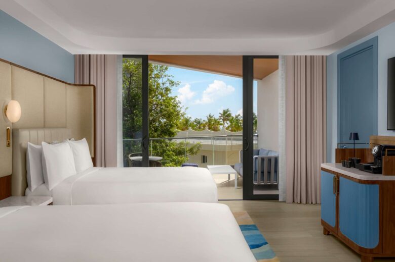 Modern hotel room with two double beds, a balcony with outdoor seating, large windows, and light-colored decor. Trees and blue sky are visible outside.