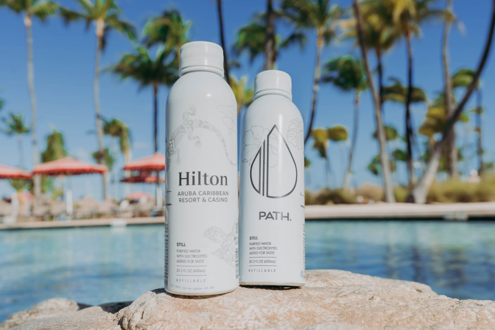 Two reusable water bottles, one labeled “Hilton Aruba Caribbean Resort & Casino” and the other “PATH,” are placed on a rock by a pool, with palm trees and red umbrellas in the background.