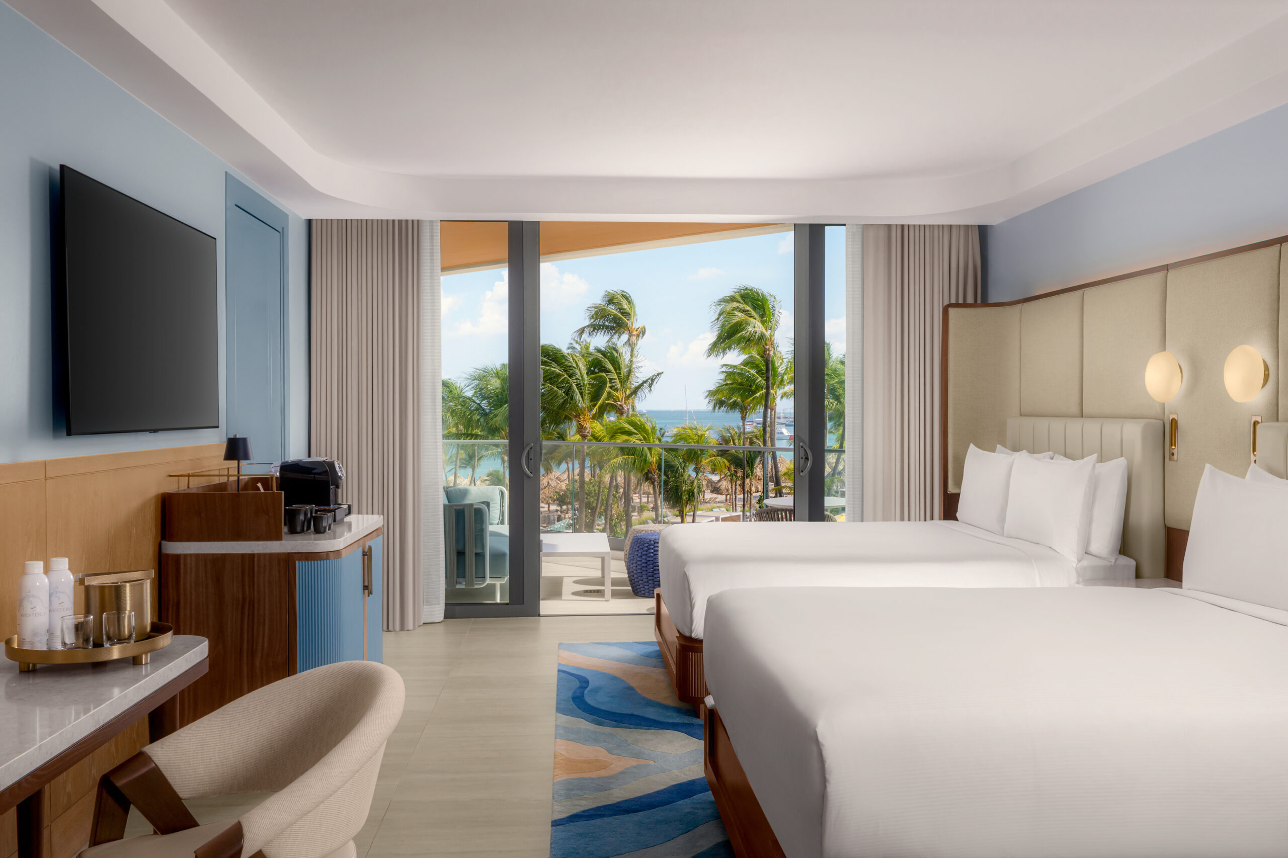 Modern hotel room with two beds, a TV, desk, and coffee station. Large window with balcony overlooks palm trees and the ocean, letting in natural light. Neutral and blue tones create a calm, relaxing atmosphere.