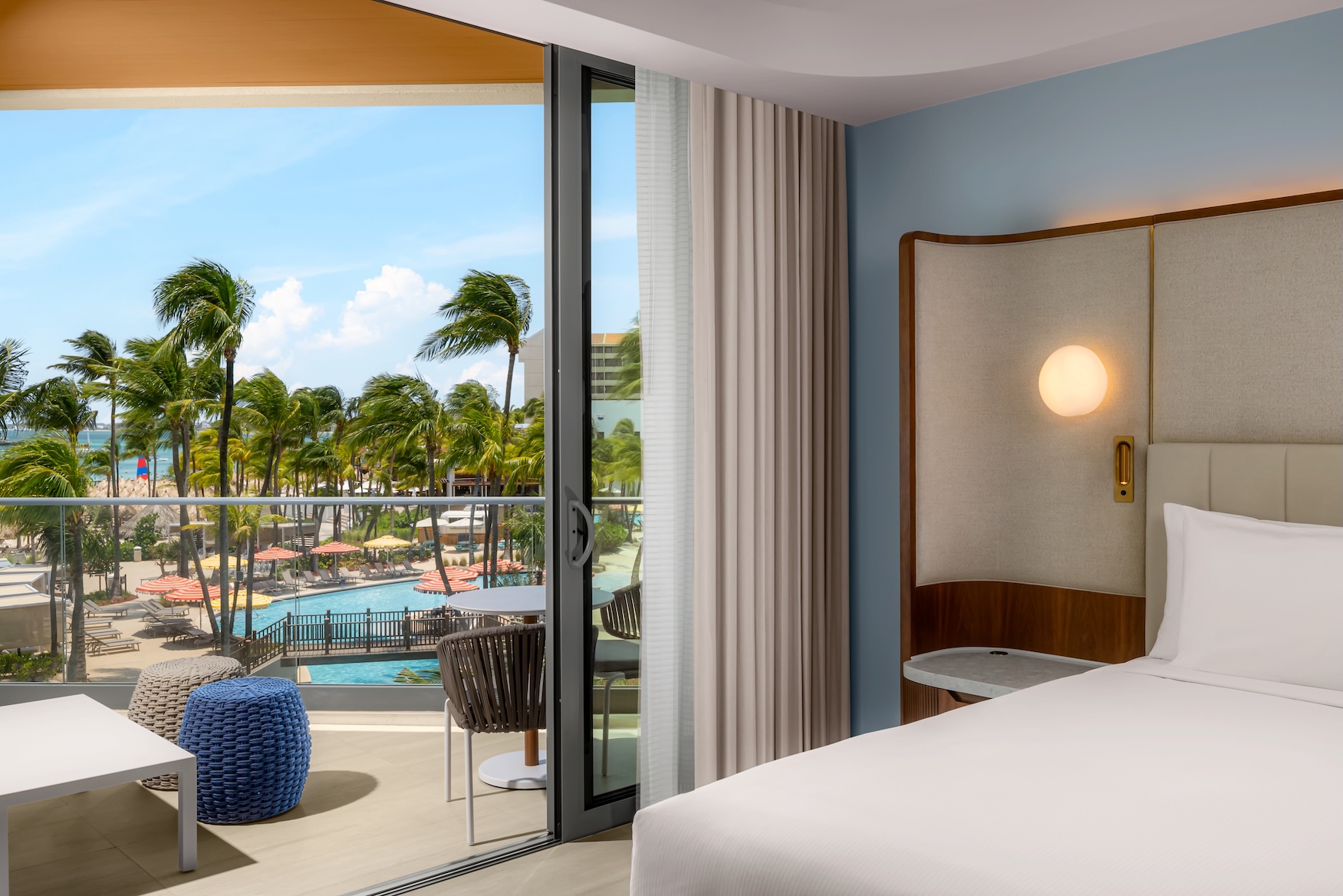 A modern hotel room with a large bed and open curtains shows a balcony view of palm trees, a pool, and the ocean under a bright blue sky.