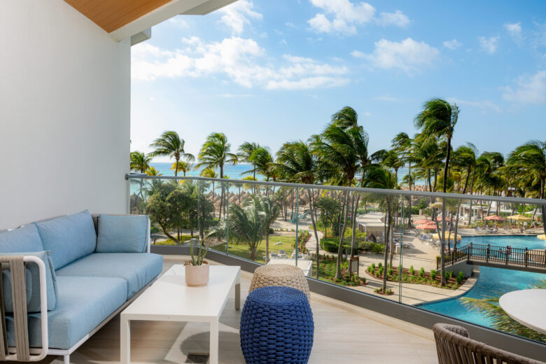 A modern balcony with a blue sofa, table, and chairs overlooks a tropical resort with palm trees, a pool, and the ocean in the background under a blue sky with scattered clouds.