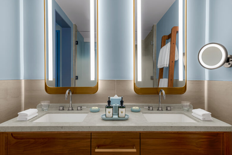 Modern bathroom with double sinks, two lighted mirrors, neatly folded towels, glass cups, and toiletries on the counter. A wooden rack with towels is visible in the background against light blue walls.