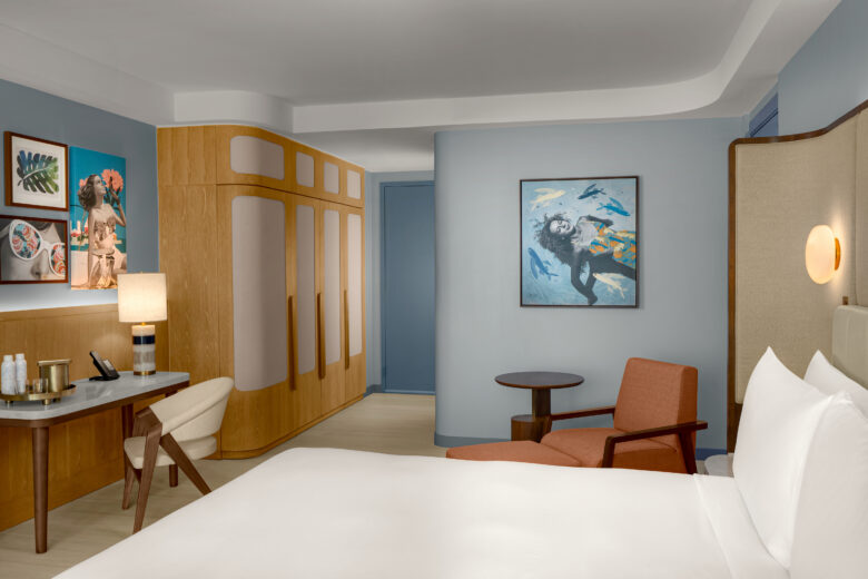 A modern hotel room with a bed, desk, chair, and artwork on blue walls. Wooden accents, a lamp, and soft lighting create a cozy, inviting atmosphere.