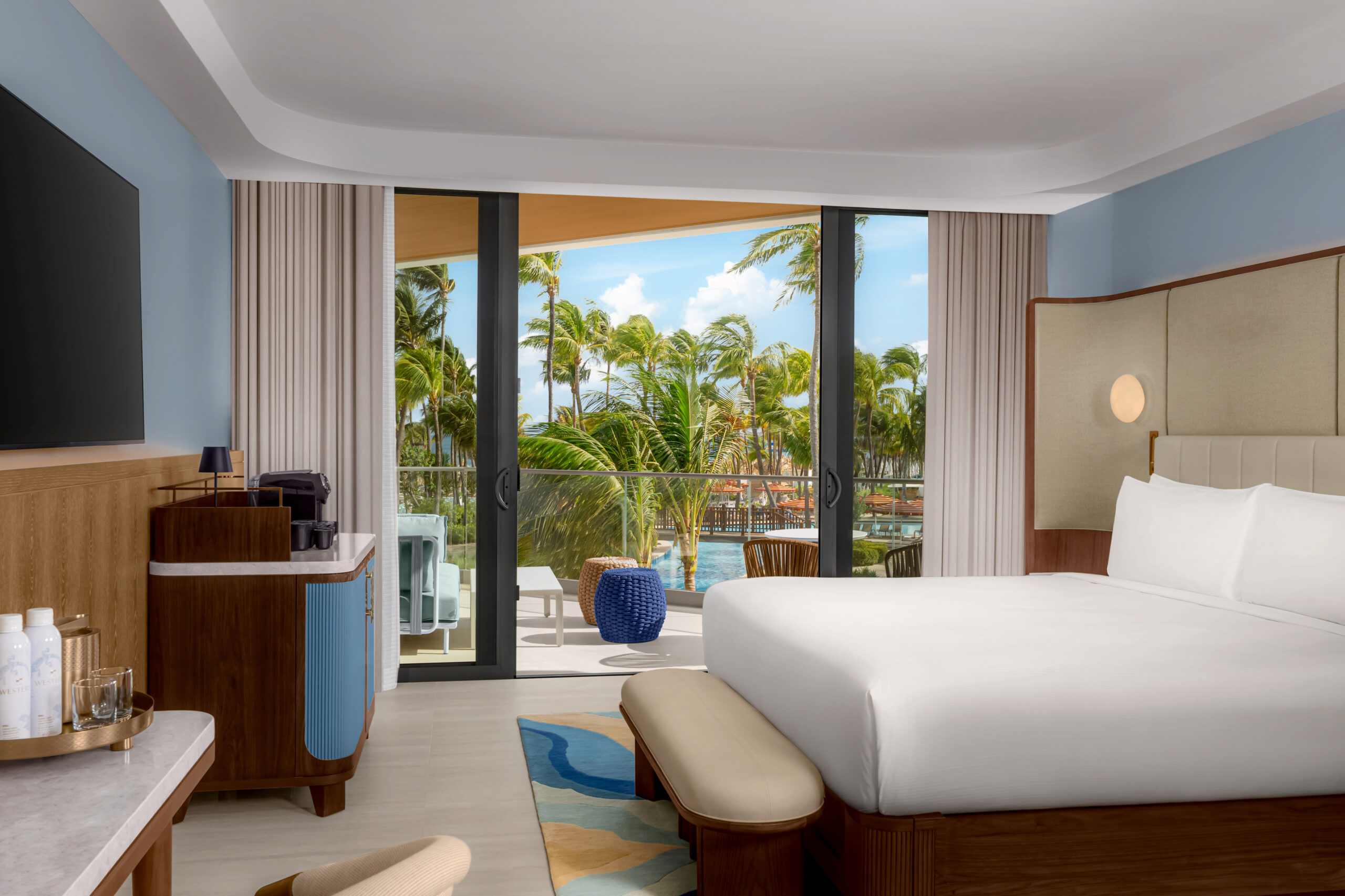 A modern hotel room with a large bed, TV, coffee station, and a balcony overlooking palm trees and a pool on a sunny day.