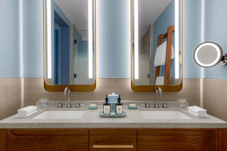 Modern bathroom with double sinks, lighted mirrors, rolled white towels, toiletries, glasses, and a wooden towel rack with folded towels against a light blue and beige wall.