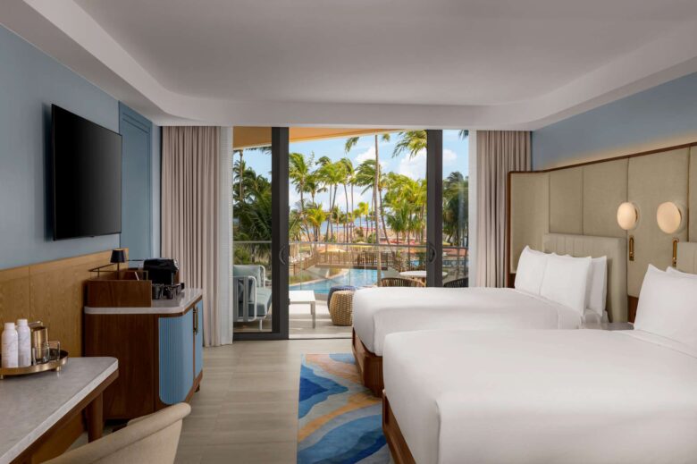 A modern hotel room with two double beds, a TV, a coffee station, and a balcony overlooking a pool and palm trees, with the ocean visible in the background.